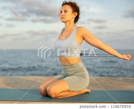 Yoga practice on embankment, female athlete fulfil carry out vajrasana with belt Yoga practice on embankment, female athlete fulfil carry out vajrasana with belt 118670938