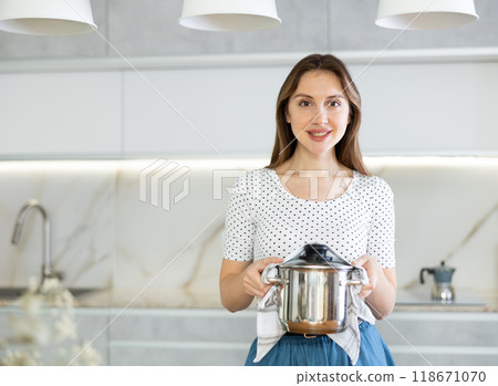 Smiling girl is standing in kitchen in casual clothes and holding stainless steel pot in hands. Smiling girl is standing in kitchen in casual clothes and holding stainless steel pot in hands. 118671070