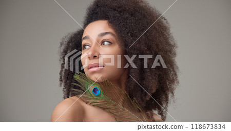 Health, beauty, tenderness and softness concept - beautiful African American woman with curly afro hairstyle and clean, healthy skin, smiles and touches her face with a bright colorful peacock feather 118673834