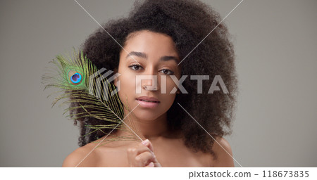 Health, beauty, tenderness and softness concept - beautiful African American woman with curly afro hairstyle and clean, healthy skin, smiles and touches her face with a bright colorful peacock feather 118673835