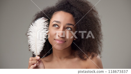 Health, beauty, tenderness and softness concept - beautiful African American woman with curly afro hairstyle and clean, healthy skin, smiles and touches her face with a white fluffy feather 118673837