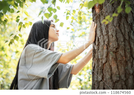 A young woman touches the bark of a tree while standing in a sunlit forest  118674560
