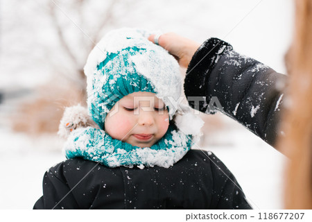 Boy child in a turquoise hat and scarf is covered in snow. Mom touches the baby's head 118677207