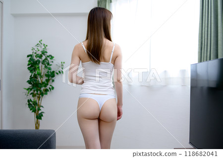 A young, slender woman in underwear with a beautiful back view walks through a room by the window, bathed in the morning sun 118682041
