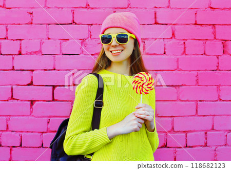 happy smiling teenager girl with colorful lollipop wearing yellow sweater, hat on pink background 118682123
