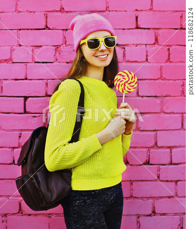 happy smiling teenager girl with colorful lollipop wearing yellow sweater, hat on pink background 118682124