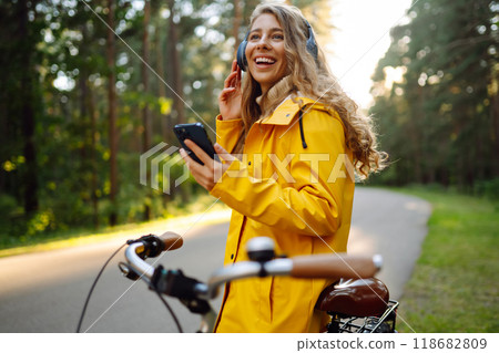 Woman using mobile phone in city while listening music through earphones to go by bicycle in park. Woman using mobile phone in city while listening music through earphones to go by bicycle in park. 118682809