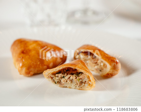 Small russian pies with minced tuna fish Russian Piroshki. Cooked tuna fish hand portioned pies or empanadas cut in half on white plate in elegant restaurant interior. Selective focus, close up. 118683806