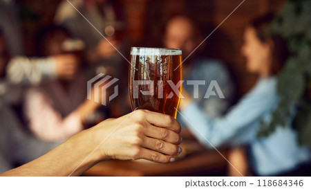 Raised glass of beer in focus, while group of friends in blurred background cheer and celebrate, likely in cozy pub. Emotions joyful and festive. Raised glass of beer in focus, while group of friends in blurred background cheer and celebrate, likely in cozy pub. Emotions joyful and festive. 118684346