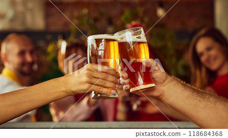Close-up of two raised glasses filled with amber beer, with blurry background action suggesting lively sports crowd enjoying game together. 118684368