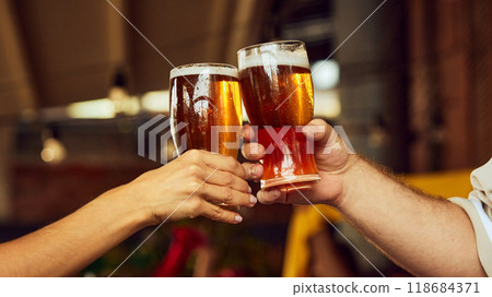Two hands clink glasses, foam spilling over. Blurred cheering fans in background elevate celebratory spirit. Meeting with old friends. 118684371