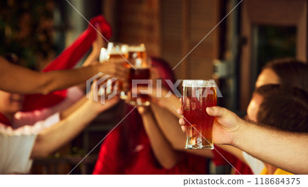 Two glasses clink as sport fans toast. Blurred figures in background add depth to scene of joyful cheers. Selective focus. Meeting with old friends. 118684375