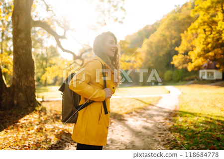 A smiling tourist in a bright coat walks through an autumn park in sunny weather. Travel concept. 118684778