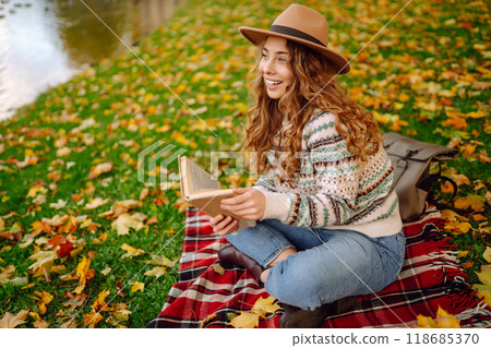 Beautiful curly woman in hat on mat with book in autumn park. Relaxation, solitude with nature. Beautiful curly woman in hat on mat with book in autumn park. Relaxation, solitude with nature. 118685370