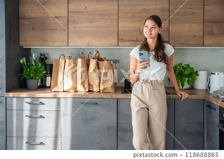 Young woman with smart phone in hand next to paper bags full of food in kitchen. Groceries ordered online and delivered by courier 118688865