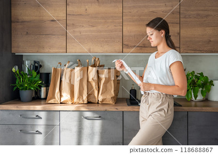 Young woman with receipt from the store next to paper bags full of food in kitchen. Groceries ordered online and delivered by courier 118688867