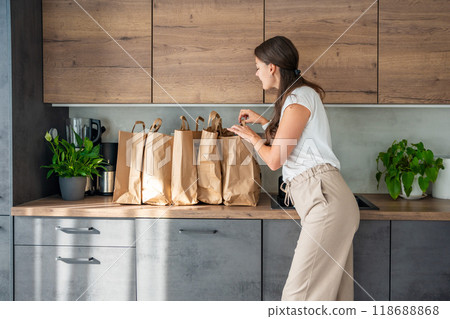 Young woman next to paper bags full of food in kitchen. Groceries ordered online and delivered by courier Young woman next to paper bags full of food in kitchen. Groceries ordered online and delivered by courier 118688868