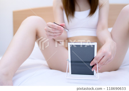 VIO精緻區 一位年輕女子在自我照顧自己的胯部,一邊照鏡子一邊用鑷子拔出鑷子,日光白色 VIO精緻區 一位年輕女子在自我照顧自己的胯部,一邊照鏡子一邊用鑷子拔出鑷子,日光白色 118690633