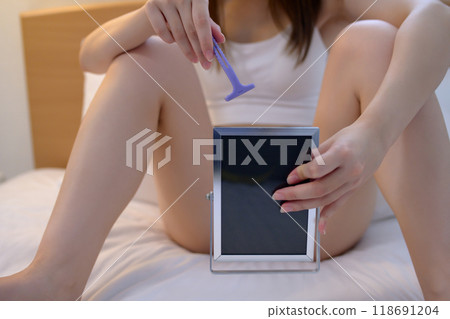 VIO精緻區一位年輕女子自我照顧自己的胯部，一邊照鏡子一邊用T型刮鬍刀剃須，脫毛，深色 118691204