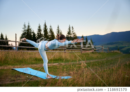 Woman practicing yoga outdoors in the mountains in a serene, natural setting. Female performing yoga pose on blue mat, with backdrop of beautiful mountain landscape at sunrise or sunset. Woman practicing yoga outdoors in the mountains in a serene, natural setting. Female performing yoga pose on blue mat, with backdrop of beautiful mountain landscape at sunrise or sunset. 118691570