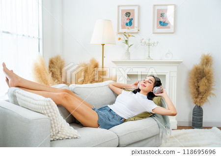 Wide shot of beautiful Asian woman lie on sofa in living room of her house enjoy to listen song with earphone with happiness. 118695696