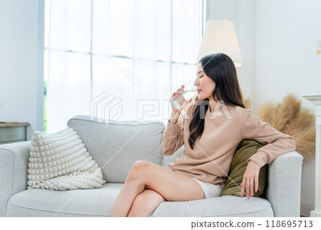 Young beautiful Asian woman sit on sofa and drink water in glass and in living room of her house. Young beautiful Asian woman sit on sofa and drink water in glass and in living room of her house. 118695713