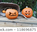 Two jack-o-lanterns with scary faces stand next to a rustic basket on a wooden table against a background of lush foliage. 118696875