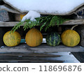 A rustic winter scene with pumpkins lined up under a snow-laden roof adorned by evergreen foliage. 118696876