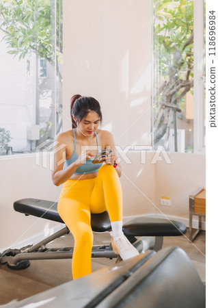 Lazy woman in Yellow Blue fitness outfit sitting and using mobile in the gym. 118696984