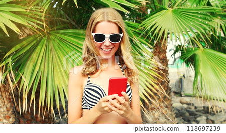 Summer portrait of happy smiling young woman with smartphone on the beach on palm tree background Summer portrait of happy smiling young woman with smartphone on the beach on palm tree background 118697239