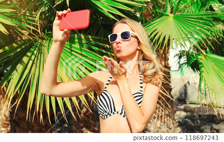 Beautiful young woman taking selfie with smartphone blowing kiss on beach on palm tree background 118697243