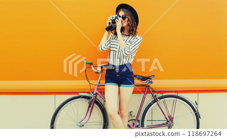happy young woman photographer taking a picture by camera with bicycle in city on orange background 118697264