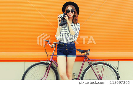happy young woman photographer taking a picture by camera with bicycle in city on orange background 118697265