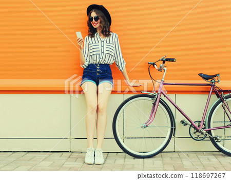 Summer colorful portrait of happy smiling young woman with smartphone, bicycle on orange background 118697267