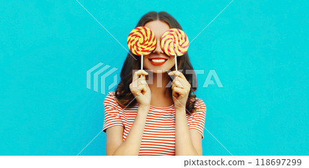 Portrait of happy smiling young woman covering her eyes with lollipop on blue background Portrait of happy smiling young woman covering her eyes with lollipop on blue background 118697299