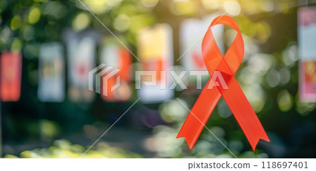 Red cancer awareness ribbon with posters in the background in an outdoor setting,  with blurred greenery and sunlight Red cancer awareness ribbon with posters in the background in an outdoor setting,  with blurred greenery and sunlight 118697401