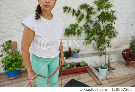 Woman exercising with resistance bands in a garden setting wearing casual fitness attire while surrounded by potted plants 118697503
