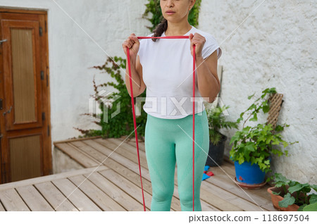 Woman practicing resistance band exercises outdoors on a wooden patio surrounded by greenery in a peaceful setting 118697504