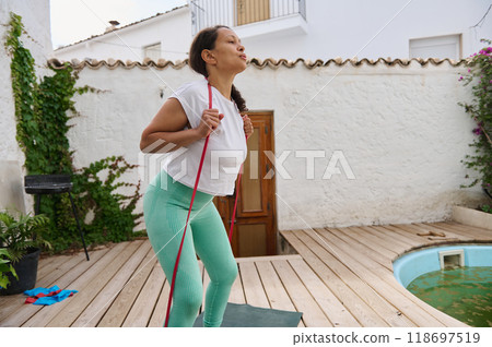 Young woman exercising with resistance band outdoors in a charming courtyard setting, wearing vibrant athletic wear, promoting fitness, wellness, and healthy lifestyle. 118697519