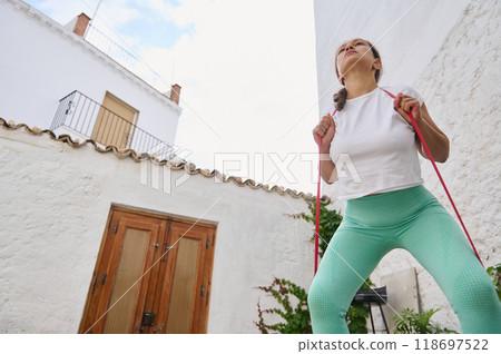 Woman exercising outdoors with resistance bands, promoting a healthy lifestyle in a courtyard setting under a clear blue sky 118697522
