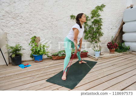 Woman practicing resistance band workout at home on a wooden deck surrounded by plants for outdoor fitness and relaxation 118697573