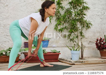 Woman practicing yoga with resistance band outdoors, focusing on flexibility and strength in peaceful garden setting 118697574