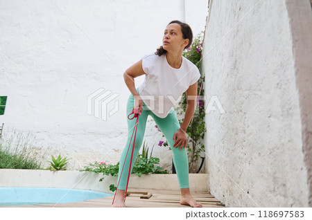 Woman in activewear exercising with resistance band near a pool outdoor on a sunny day, focused on fitness and wellness 118697583