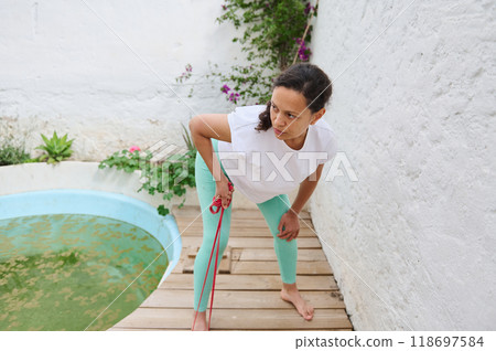 Woman exercising outdoors by a small round pool on a wooden deck, wearing white and mint activewear, focused on fitness routine Woman exercising outdoors by a small round pool on a wooden deck, wearing white and mint activewear, focused on fitness routine 118697584