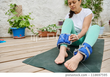 Woman preparing for outdoor yoga by adjusting ankle weights, enjoying a serene workout session on a wooden deck surrounded by plants. Woman preparing for outdoor yoga by adjusting ankle weights, enjoying a serene workout session on a wooden deck surrounded by plants. 118697595