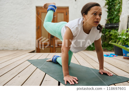 Woman practicing yoga at home with ankle weights, in a backyard setting, focusing on balance and strength on a wooden deck. Woman practicing yoga at home with ankle weights, in a backyard setting, focusing on balance and strength on a wooden deck. 118697601