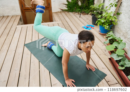 Woman practicing yoga on a mat outdoors with ankle weights, focusing on balance and strength in a peaceful garden setting 118697602