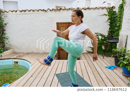 Woman practicing outdoor fitness routine on wooden deck with ankle weights near a pool in a serene setting Woman practicing outdoor fitness routine on wooden deck with ankle weights near a pool in a serene setting 118697612