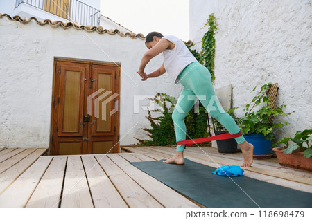 Woman practicing outdoor exercise with resistance band on a patio, improving balance and strength through home workout session in relaxed environment Woman practicing outdoor exercise with resistance band on a patio, improving balance and strength through home workout session in relaxed environment 118698479
