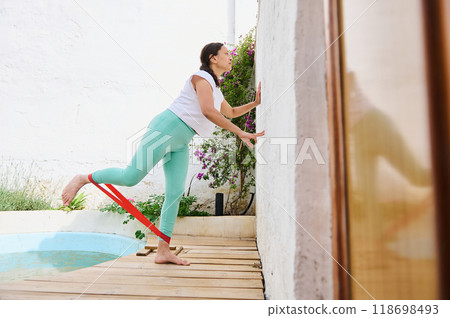 Woman practicing exercise with resistance band by a bright poolside on a sunny day, focusing on balance and strength training. 118698493